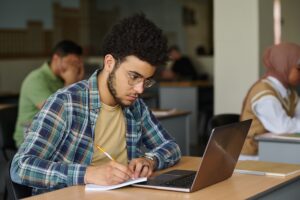 Young man sitting at desk with laptop and making notes in notepad, he learning language in class