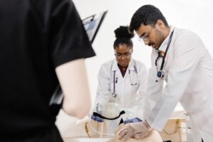 Two medical students in white coats practice CPR on a mannequin while a third student observes.