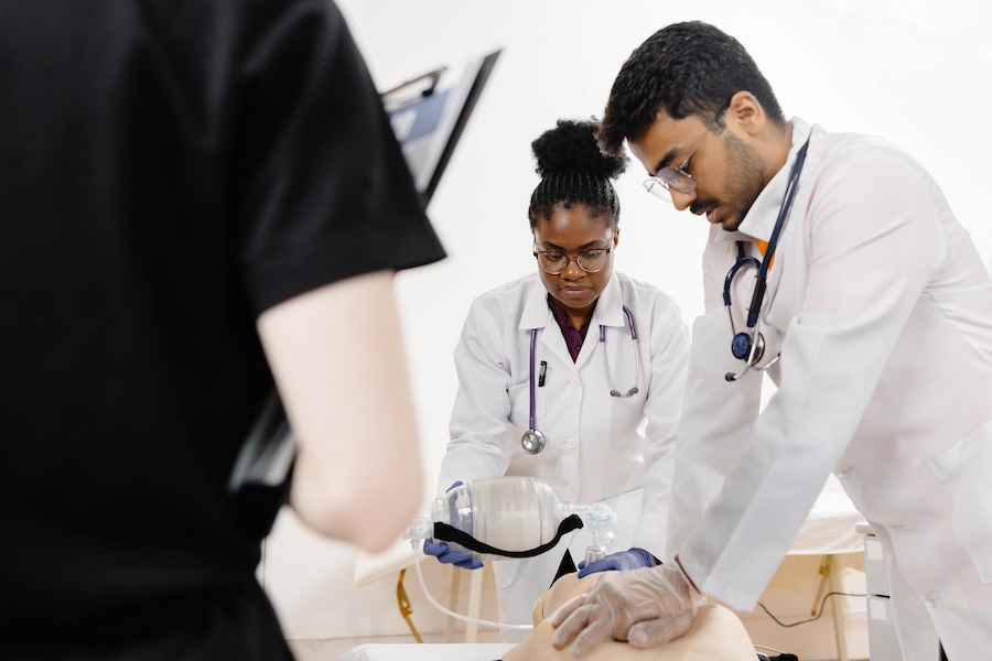 Two medical students in white coats practice CPR on a mannequin while a third student observes.