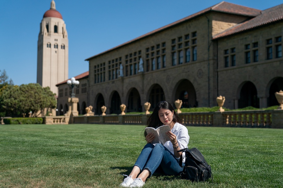 leisure asian female exchange student enjoying reading while sitting on green lawn in front of hoover tower during break time on a sunny day at school university in California usa