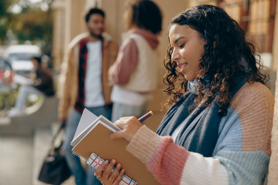 Young female student studying while standing outside of university on friends background