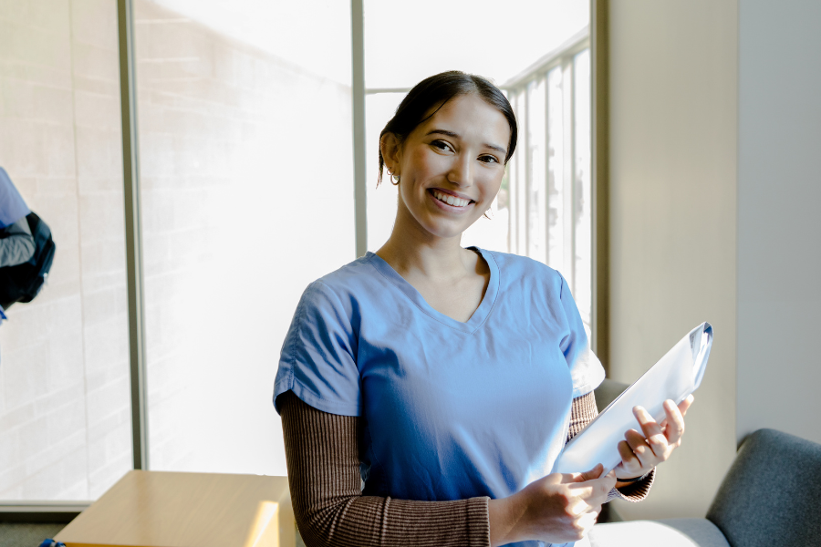 Smiling pre-med student in blue scrubs holding study materials, standing in a bright room, symbolizing preparation and success in medical school