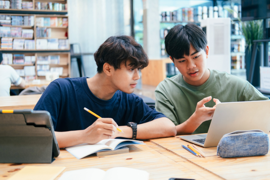 Two high school students working together on academics, discussing notes and using a laptop for study resources in a library setting.