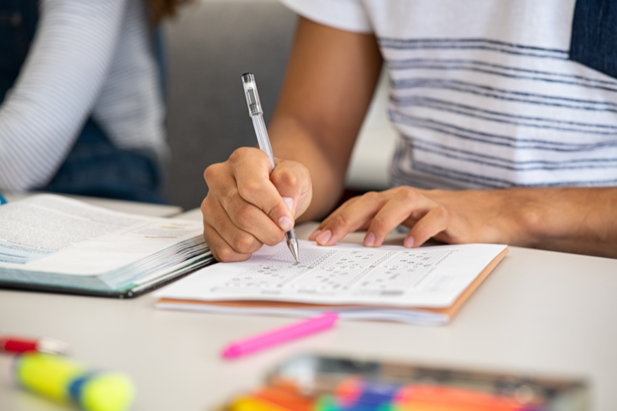 Student completing a test preparation workbook with a pen, surrounded by study materials, highlighting the focus and effort involved in test prep.