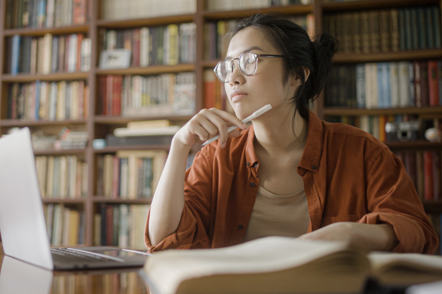 A thoughtful student sitting at a desk in a library, holding a pen and reflecting while studying, representing deep thinking for college essays.